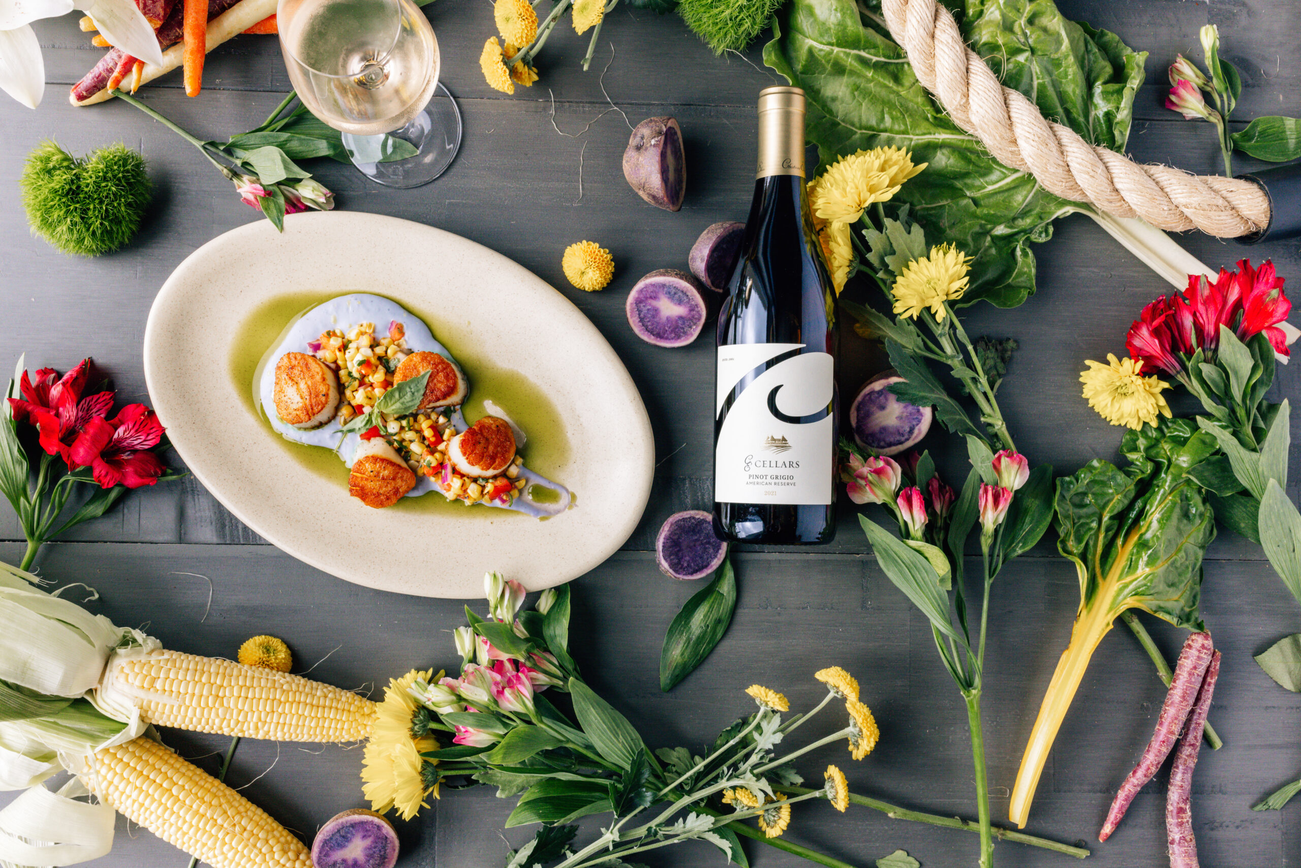 A flat lay photo of a plate of scallops, a bottle of wine, and various vegetables and flowers on a dark surface.