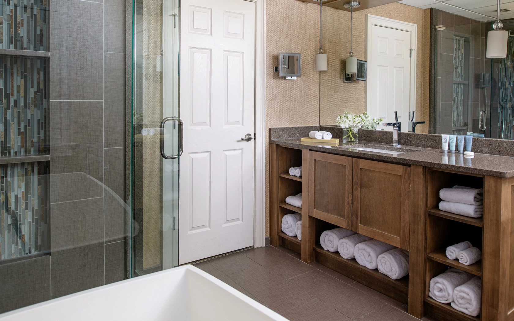 Spa bathroom with glass walk-in shower, teal mosaic tile accent wall, wood vanity with open shelving, stacked white towels, and freestanding soaking tub