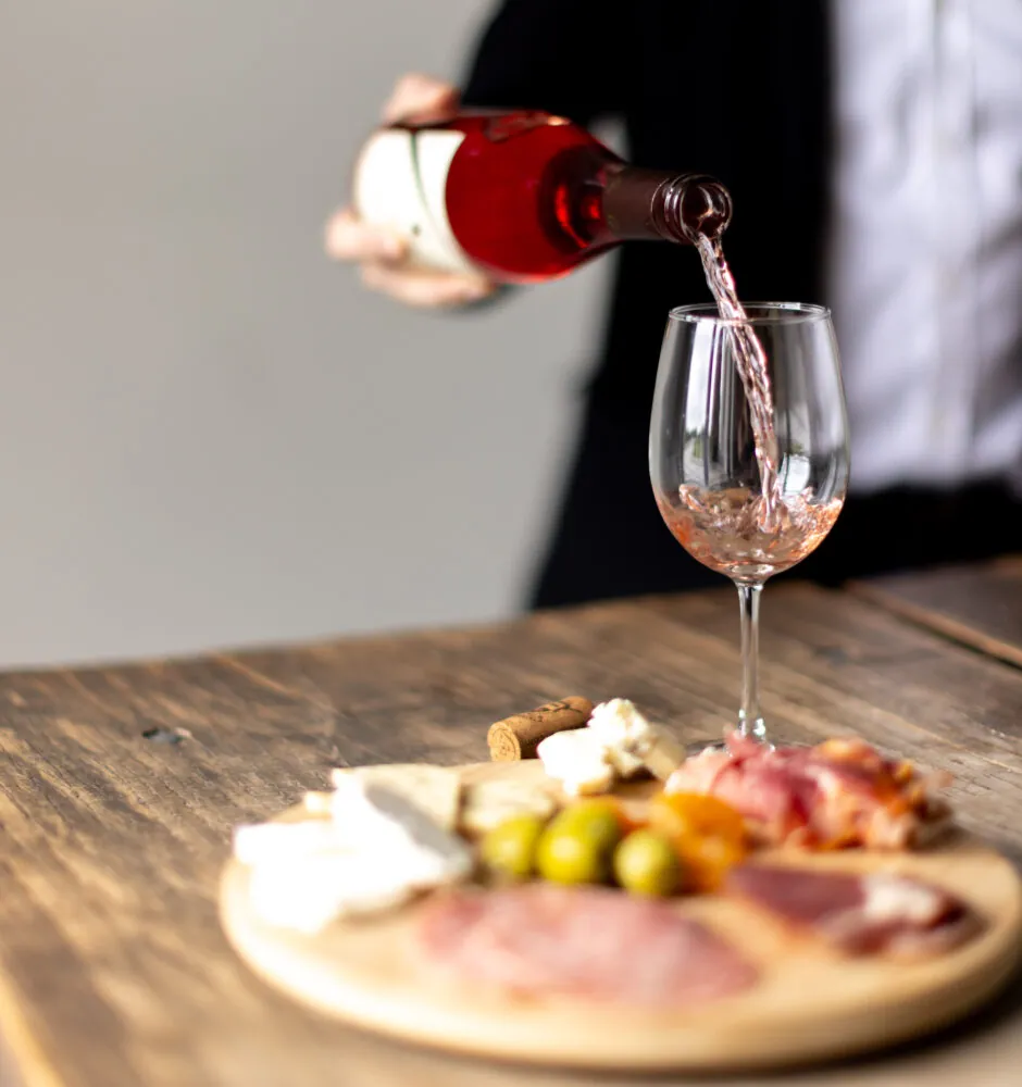 A close-up, blurry shot shows a person in a dark jacket and white shirt pouring rosé wine into a glass. In the foreground, there is a round wooden board with an assortment of snacks, including meats, olives, and cheese.