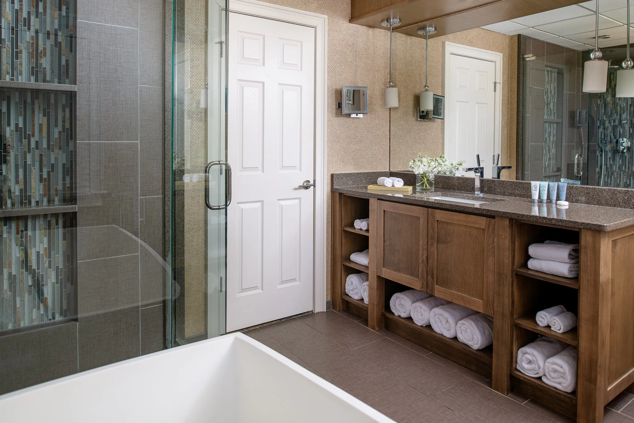 Spa-inspired bathroom with glass-enclosed walk-in shower featuring teal mosaic tile, wood vanity with open shelving, stacked white towels, and freestanding soaking tub