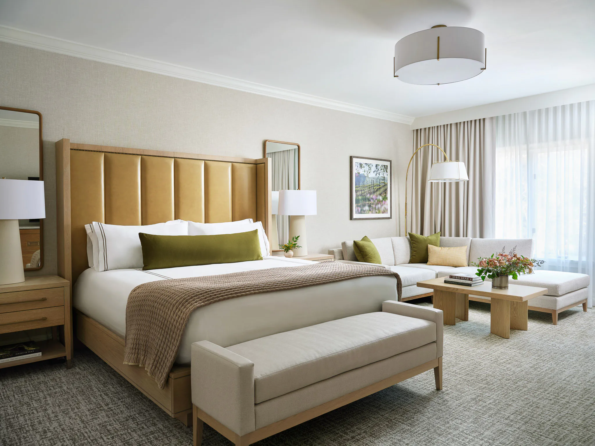 Spacious King room with amber paneled headboard, olive green pillows, grey upholstered bench, sofa seating area, and arched window with natural light