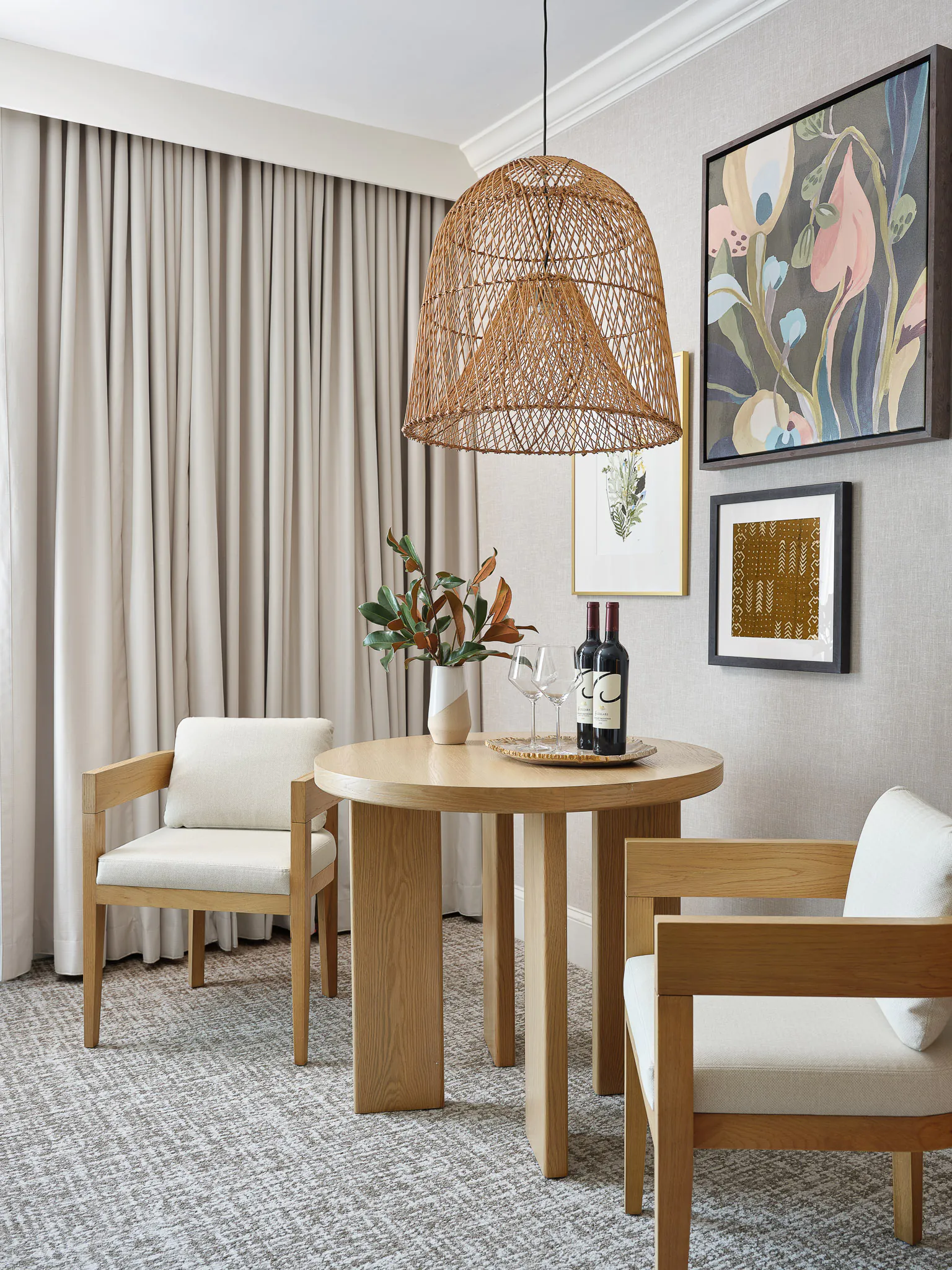 Intimate in-room dining nook with round wooden table, white chairs, rattan pendant light, bottle of wine, and colorful botanical artwork