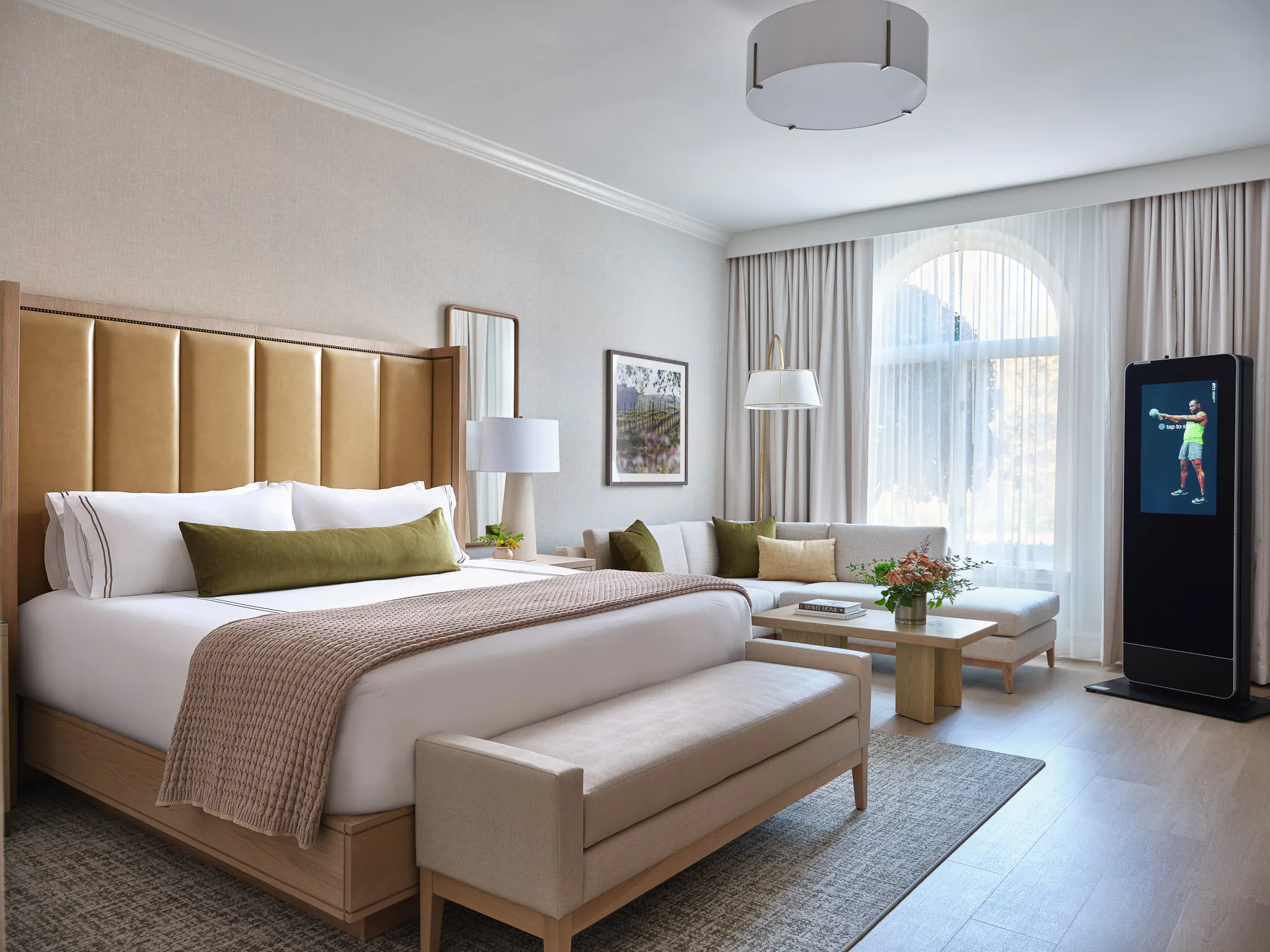 Spacious King room with amber paneled headboard, olive green pillows, grey upholstered bench, sofa seating area, and arched window with natural light