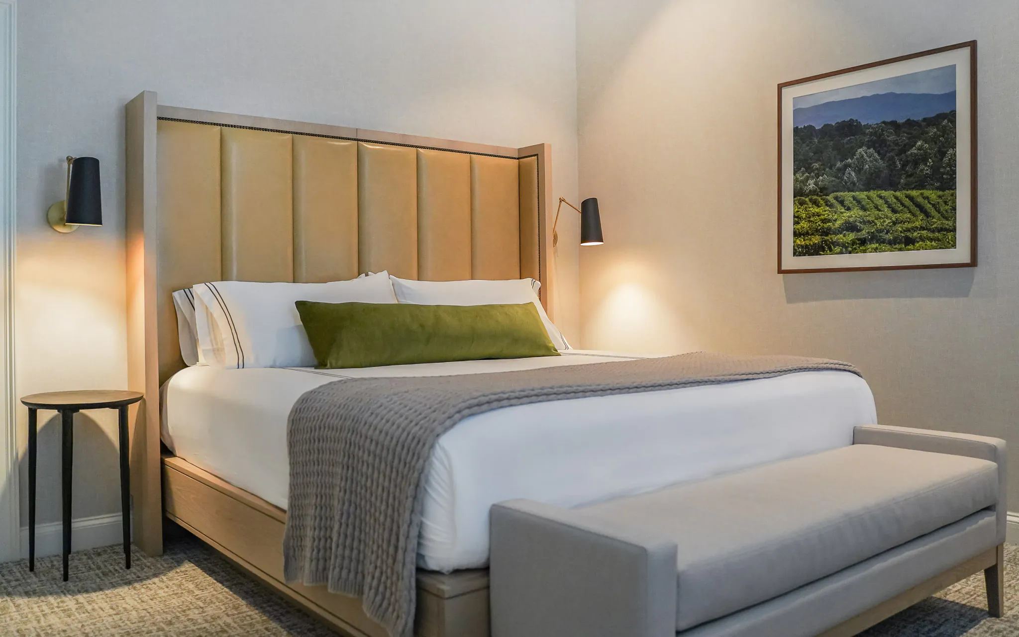 King bedroom with tall upholstered headboard, white bedding, olive green accent pillow, wall-mounted reading lights, grey upholstered bench, and framed vineyard artwork
