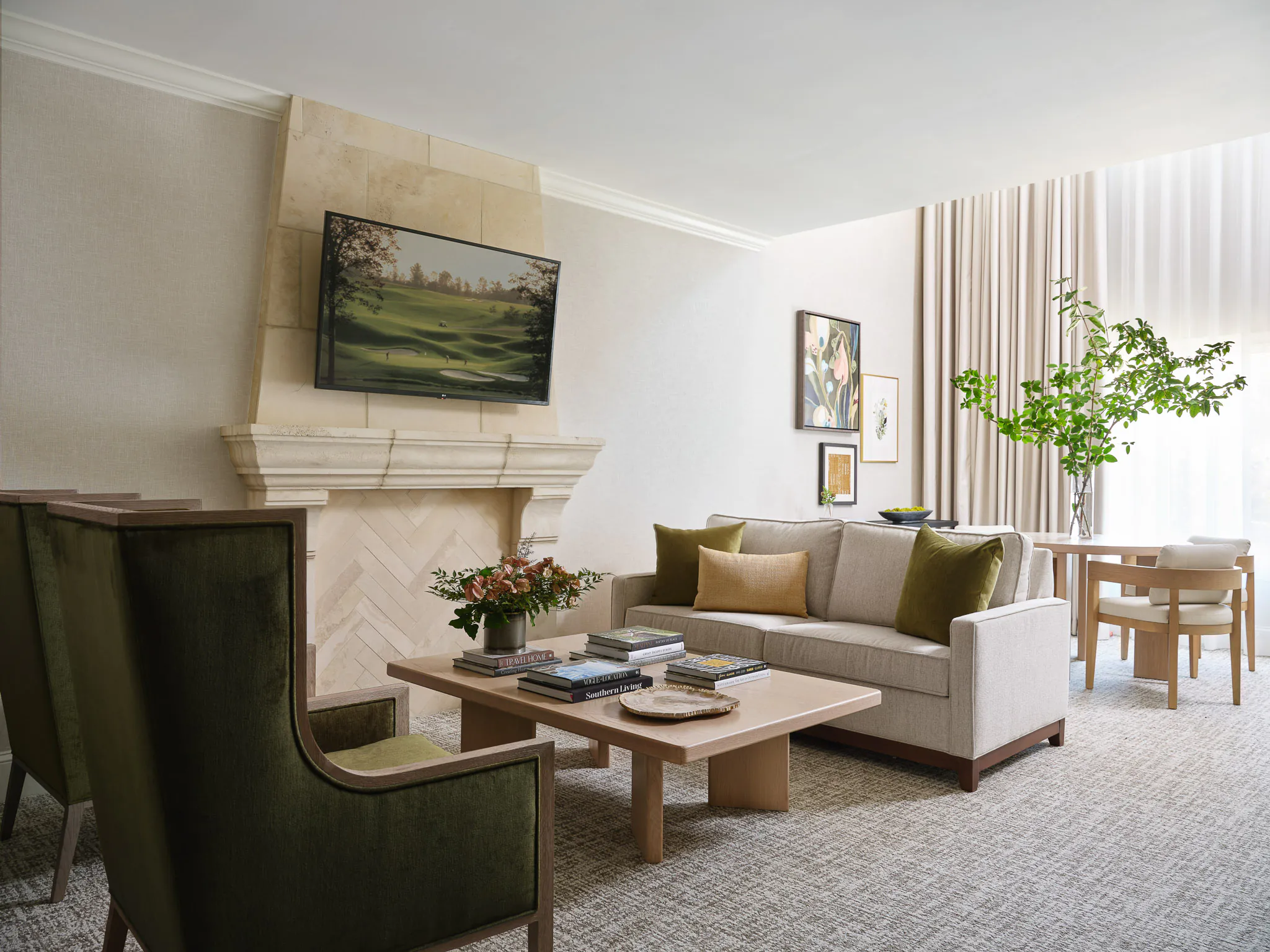 Suite living room with stone fireplace, TV, neutral sofa with olive green and gold accent pillows, dark green wingback chair, wooden coffee table, and dining area with garden views