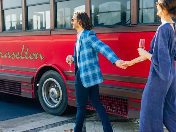 A happy couple in formal attire, holding hands and glasses of champagne, walking in front of a red bus.