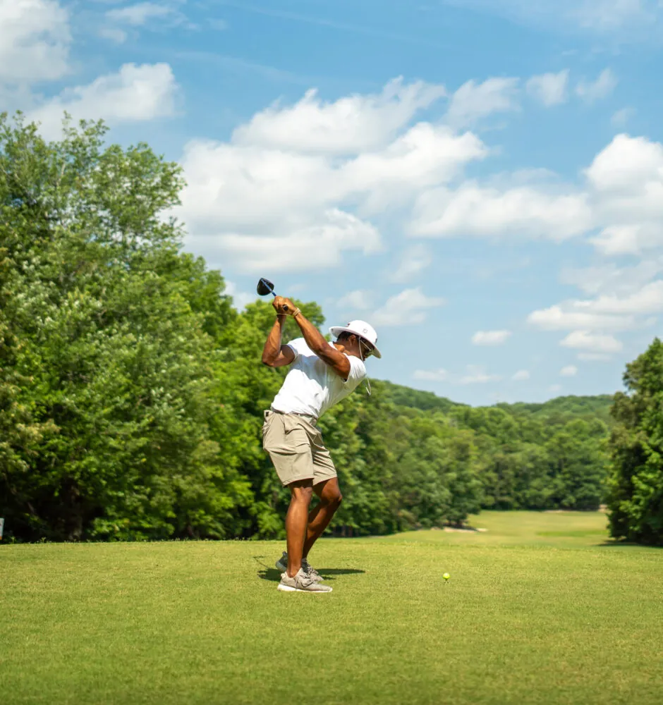 A person in a white polo shirt and shorts takes a powerful swing with a golf club on a bright, sunny day. The golf course is surrounded by trees under a blue sky with scattered clouds.