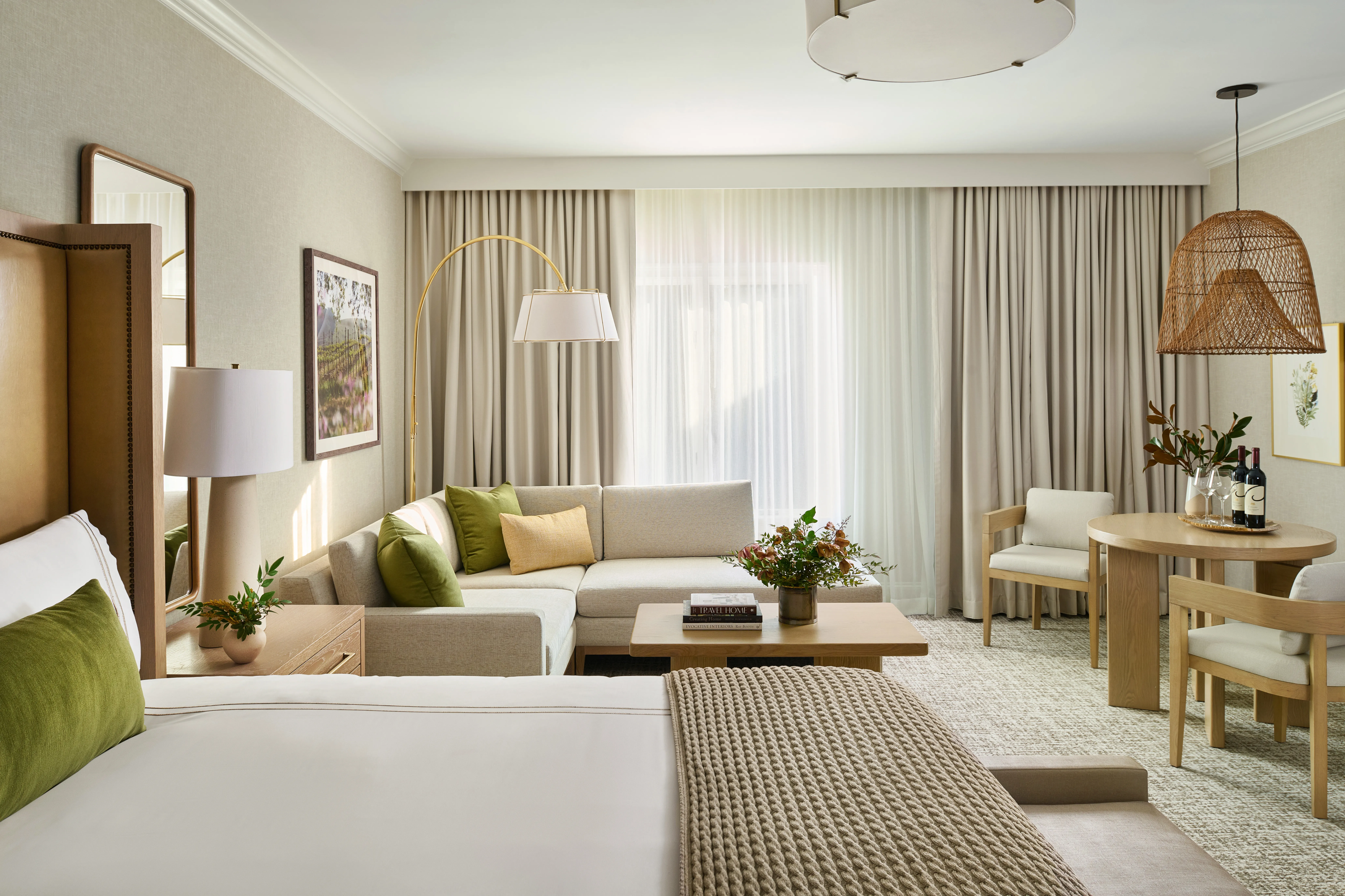 Bright suite with king bed, neutral sofa with olive green and gold pillows, wooden coffee table, rattan pendant light, and dining area with natural light