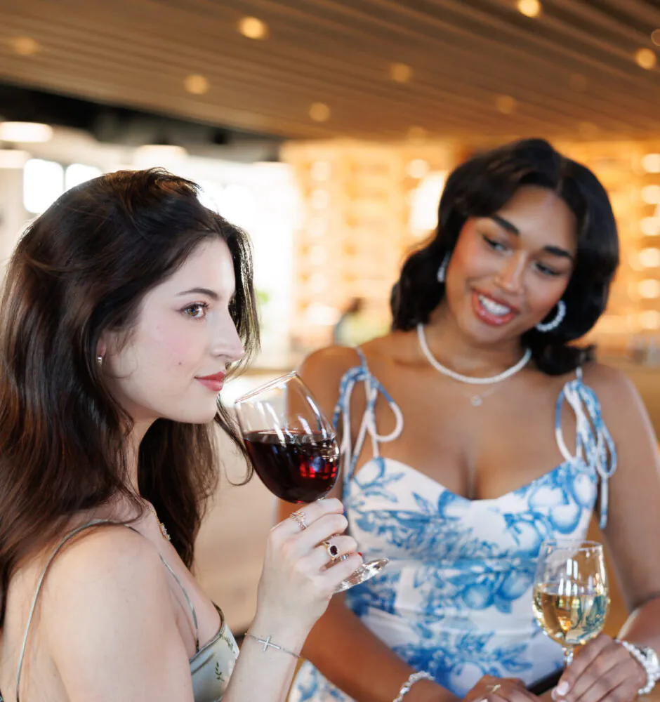 two women at a bar or counter. The woman on the left, with long dark hair, is holding a glass of red wine and looking away from the camera. The woman on the right is looking at the first woman and smiling. She is wearing a blue and white patterned dress and holding a glass of white wine. The background is a blurry indoor setting with wooden ceiling beams.