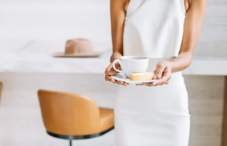 A person in a white dress holds a cup of coffee and a saucer.