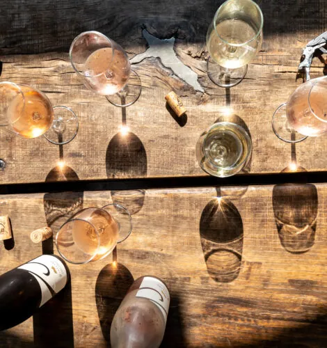 An overhead, blurred shot of a rustic wooden surface with wine glasses, two wine bottles, and corks. The glasses cast long shadows and contain a light-colored wine, possibly white or rosé. A corkscrew is also visible near the top of the image.