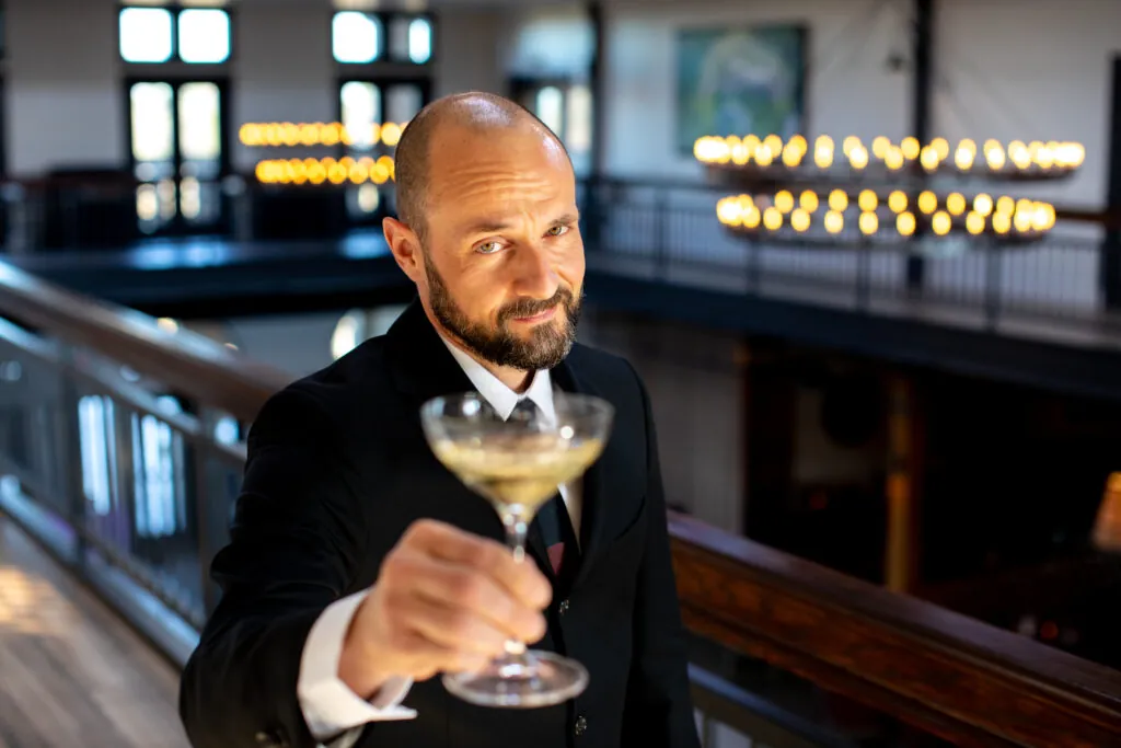 A man holds up a glass with a clear drink and an olive in it.