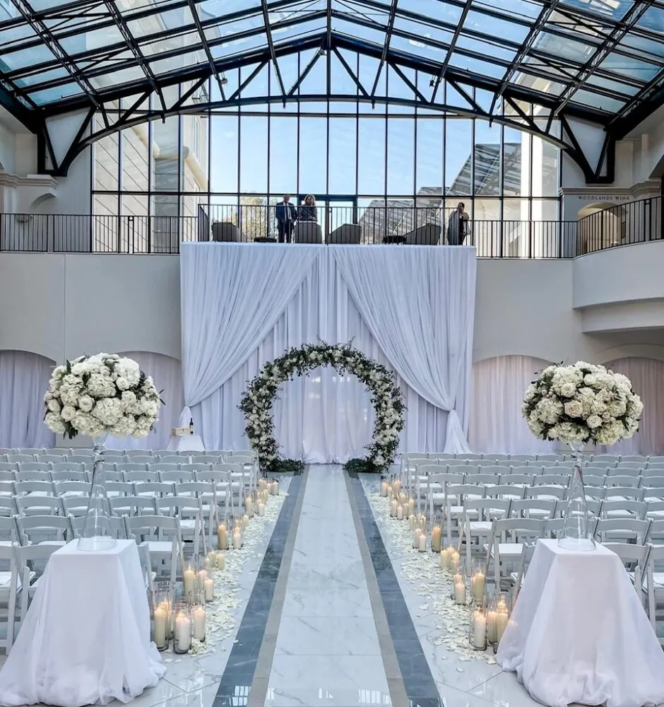Indoor wedding ceremony with white chairs, a white aisle runner, and an arched floral arrangement.