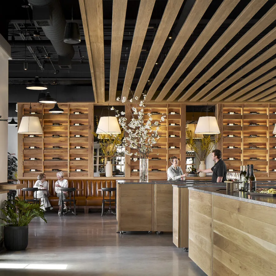 A blurred image shows the interior of a winery or restaurant. In the foreground, there is a large, light-colored wooden bar counter with a black top. Behind the counter are two people, one of whom appears to be a server talking to a customer. The back wall is lined with wooden shelves that hold wine bottles horizontally. The ceiling features parallel wooden slats.