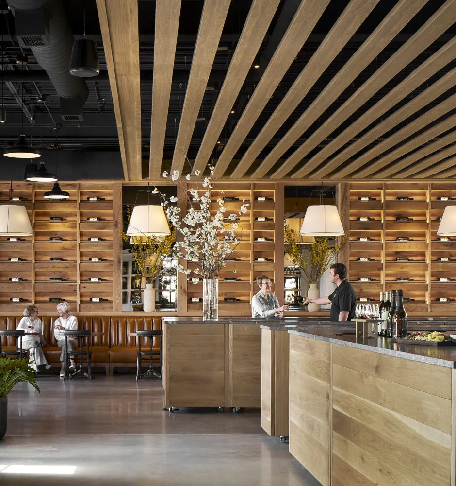 A blurred image shows the interior of a winery or restaurant. In the foreground, there is a large, light-colored wooden bar counter with a black top. Behind the counter are two people, one of whom appears to be a server talking to a customer. The back wall is lined with wooden shelves that hold wine bottles horizontally. The ceiling features parallel wooden slats.