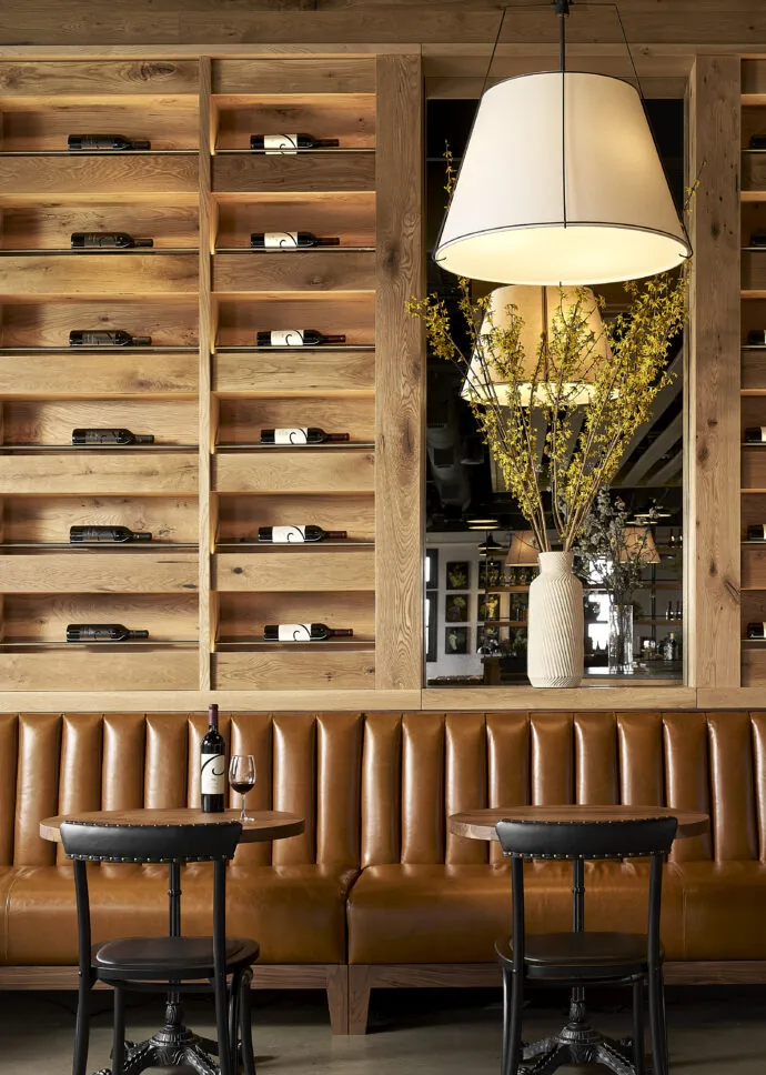 A section of the restaurant features a long, brown leather bench seat along a wall. Above the bench, there's a wall of wooden shelving holding wine bottles horizontally. A small table with two chairs is in front of the bench, and a large lamp hangs from the ceiling.