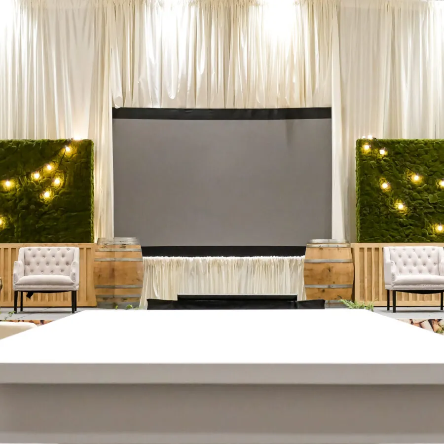 A decorated stage with a large screen, flanked by green hedges with string lights and two wooden barrels. Chairs are arranged in front of the stage.