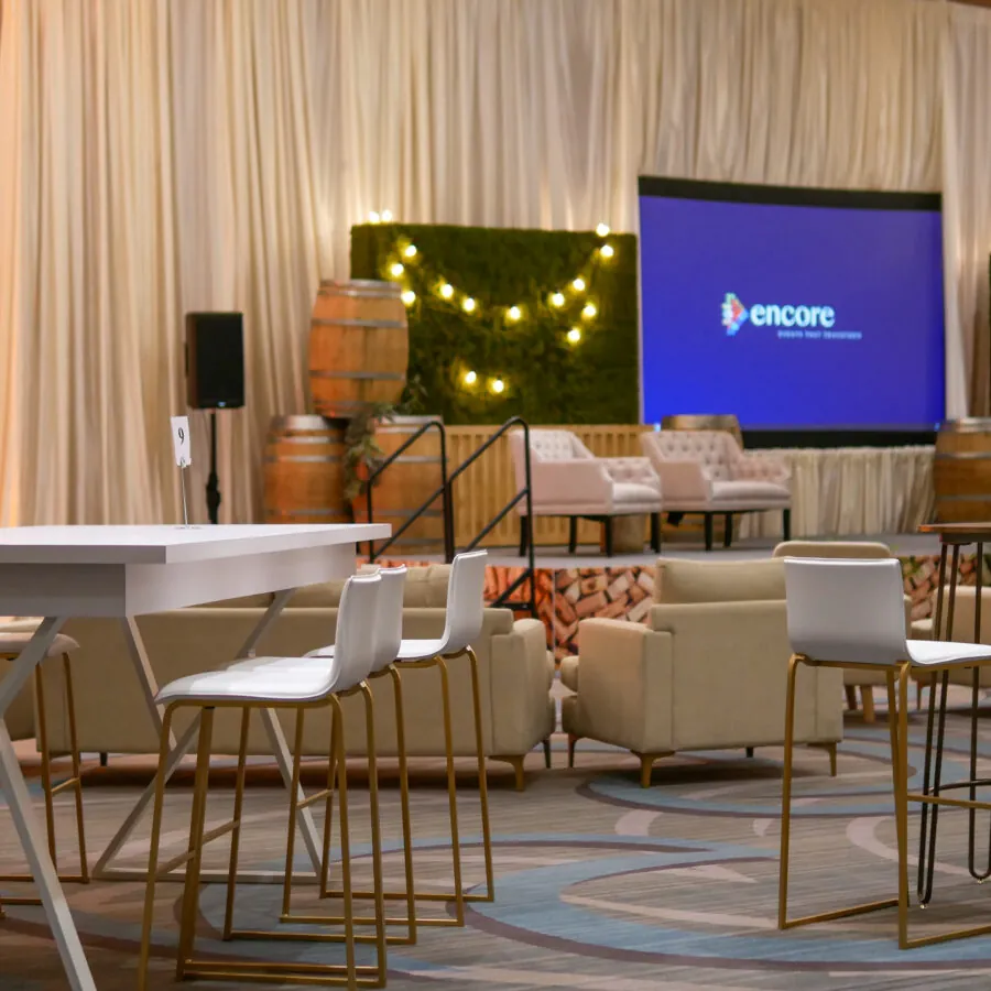 A lounge area set up for an event with couches and chairs facing a stage with a large screen, green hedges with string lights, and white curtains.