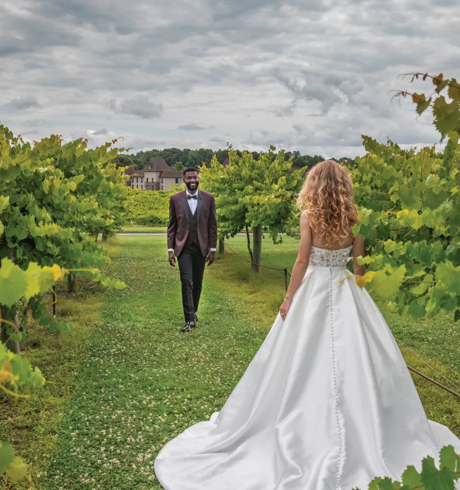 A bride in a long white dress stands in a grassy vineyard looking back at the groom, who is walking towards her.