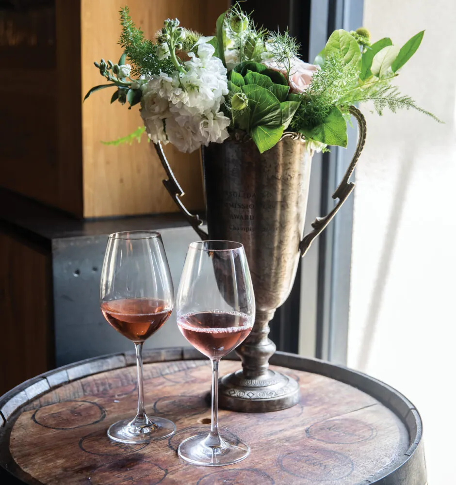 Two glasses of rosé wine on a wooden barrel, next to a flower arrangement.