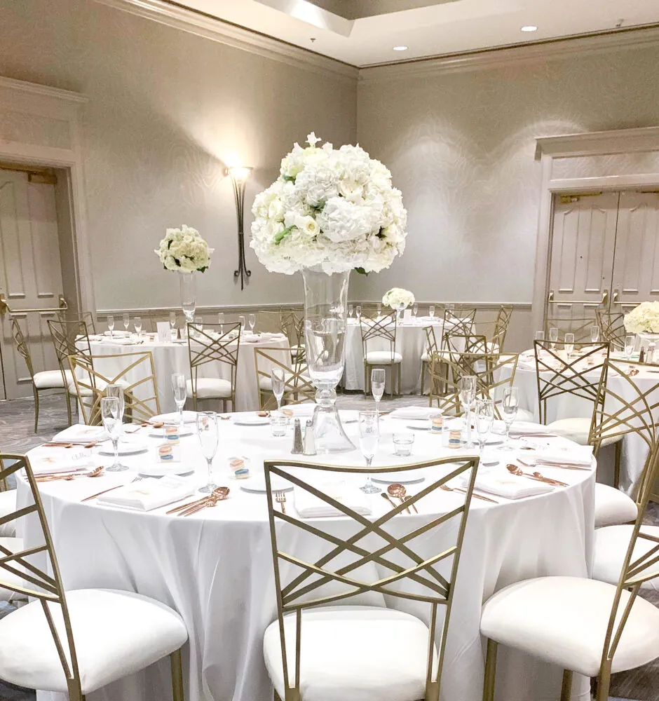 Round tables with white linens, gold chairs, and tall white floral centerpieces.