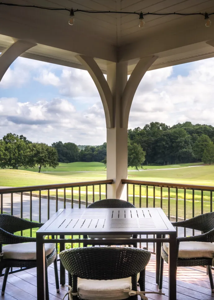 A patio with a table and chairs on a covered porch, overlooking a green golf course.
