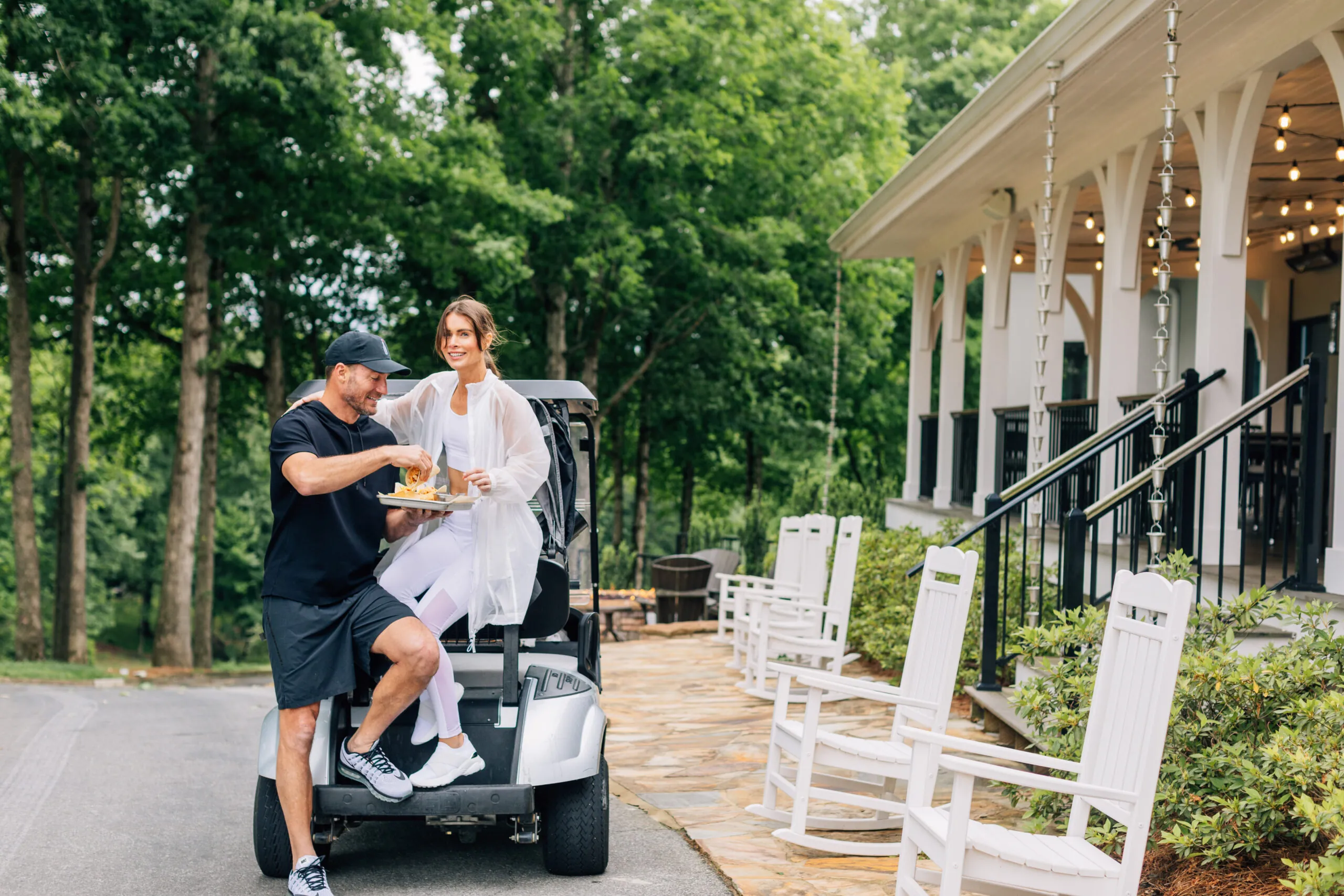 A couple on a golf cart near a building with a white porch and white rocking chairs. The man is holding a tray of drinks.