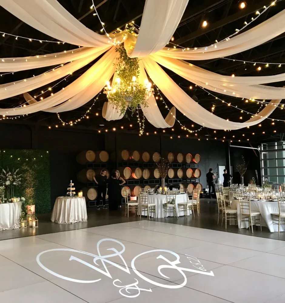 Indoor reception hall with draped fabric, string lights, and a dance floor with a monogram.