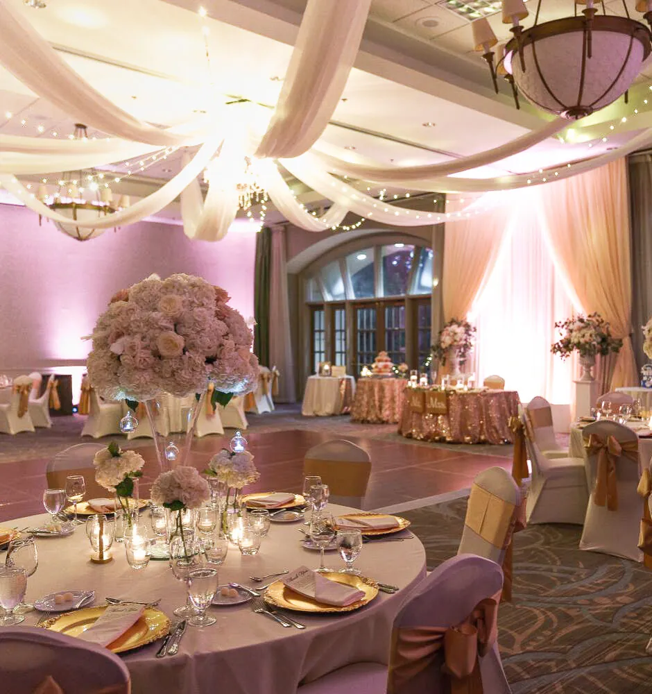 Ballroom wedding reception with draped ceilings, pink lighting, and tables decorated with gold accents and large floral centerpieces.