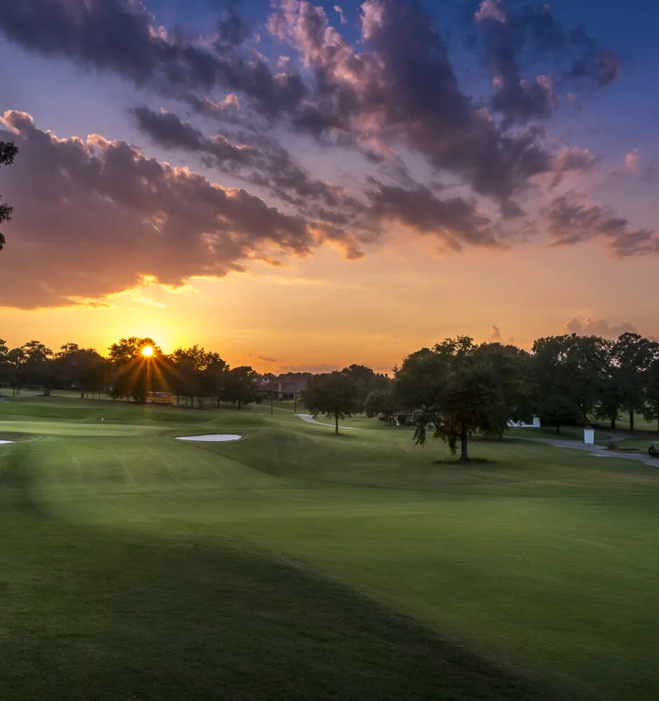A wide view of a golf course at sunset with a mix of green grass and trees. The sun is setting, creating orange and purple clouds.