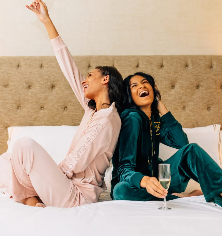 Two women in pajamas laughing on hotel bed