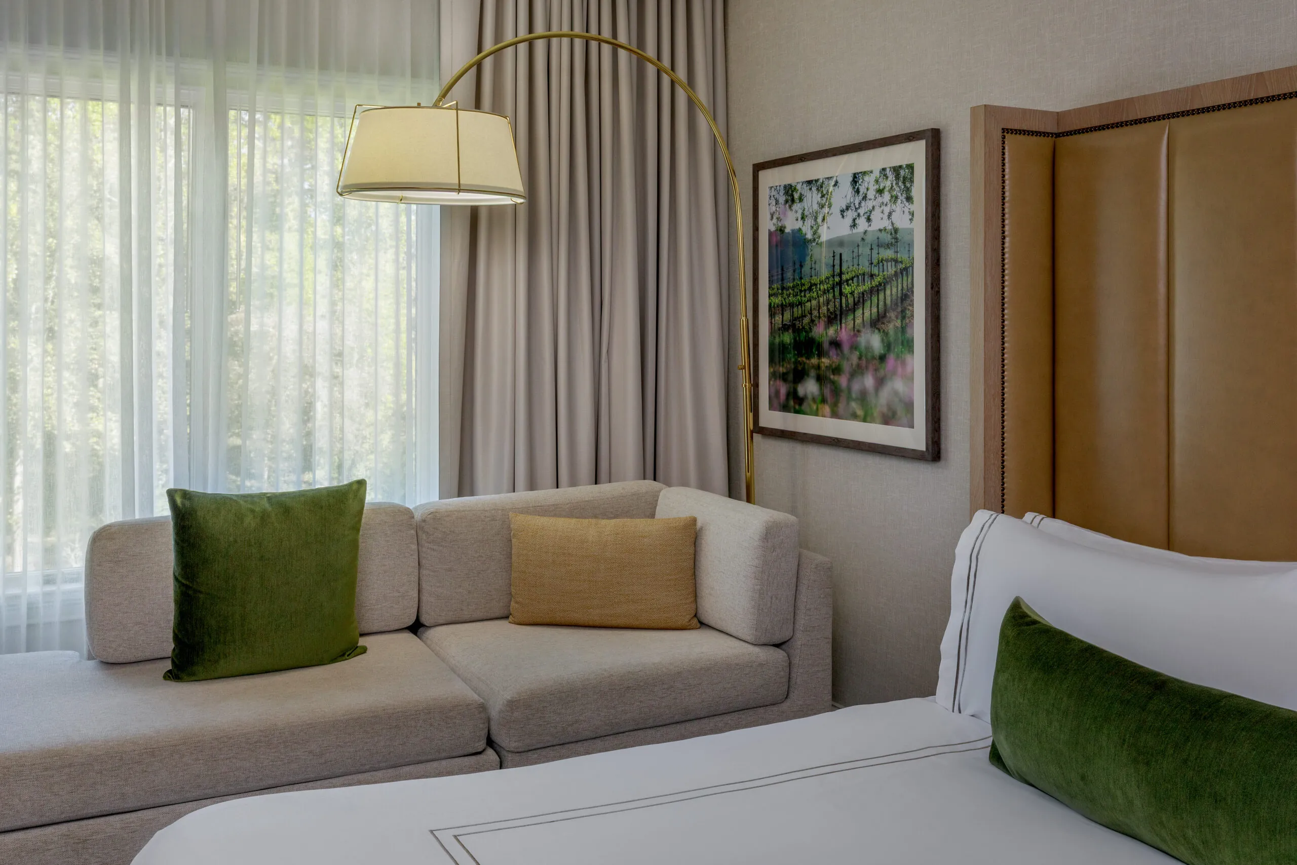 Suite seating area with grey upholstered sofa, olive green and gold accent pillows, arched floor lamp, sheer curtains, and framed botanical artwork
